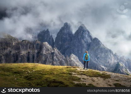 Girl with backpack on the hill looking at mountains at sunset in summer. Dramatic landscape with young woman, high rocks, trail, green grass, cloudy sky in Dolomites, Italy. Adventure and hiking