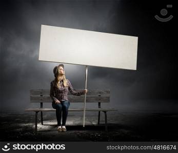 Girl with banner. Young woman in casual sitting on bench and holding white blank banner