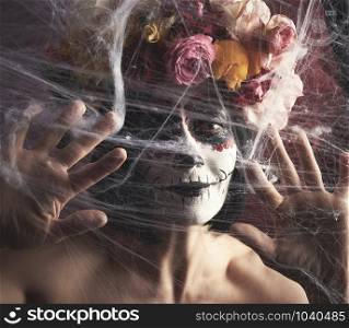 girl with black hair is dressed in a wreath of multi-colored roses and makeup is made on her face Sugar skull to the day of the Dead. Woman looking at the camera through a white web with spiders