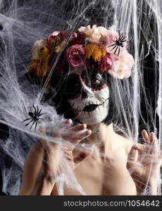 girl with black hair is dressed in a wreath of multi-colored roses and makeup is made on her face Sugar skull to the day of the Dead. Woman looking at the camera through a white web with spiders