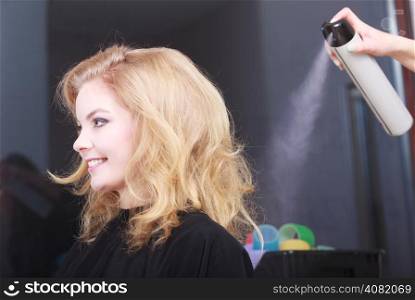 Girl with blond wavy hair by hairdresser. Hairstylist with hairspray and female client. Young woman in hairdressing beauty salon. Hairstyle.