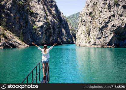 Girl with her hands up stands on bow of ship in waters of Green Canyon. In background, canyon rocks and blue sky. Concept of freedom and travel. Side, Turkey. Girl with her hands up stands on bow of ship in waters of Green Canyon