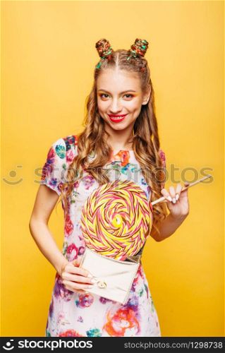 Girl with huge lollipop and little bag. Young woman gets candy caramel from her small bag. Stylish girl in colorful summer dress, yellow wall on background.. Girl with huge lollipop and little bag