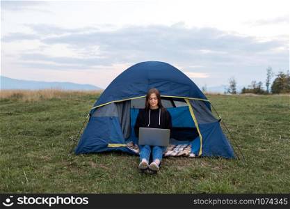 Girl with laptop in the mountains. Tourist woman uses a laptop in nature, freelancer typing on a computer near a tent in a journey against the backdrop of a mountain landscape.. Girl with laptop in the mountains. Tourist woman uses a laptop in nature, freelancer typing on a computer near a tent in a journey against the backdrop of a mountain landscape
