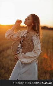 girl with long hair and straw bag in hand in the summer at sunset in the field for a walk. she is happy, her eyes are closed. background blurred art photography.. girl with long hair and straw bag in hand in the summer at sunset in the field for a walk. she is happy, her eyes are closed. background blurred art photography