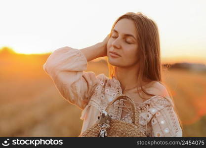 girl with long hair and straw bag in hand in the summer at sunset in the field for a walk. she is happy, her eyes are closed. background blurred art photography.. girl with long hair and straw bag in hand in the summer at sunset in the field for a walk. she is happy, her eyes are closed. background blurred art photography