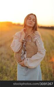 girl with long hair and straw bag in hand in the summer at sunset in the field for a walk. she is happy, her eyes are closed. background blurred art photography.. girl with long hair and straw bag in hand in the summer at sunset in the field for a walk. she is happy, her eyes are closed. background blurred art photography