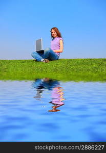 Girl with notebook sitting on grass against sky with reflection on water