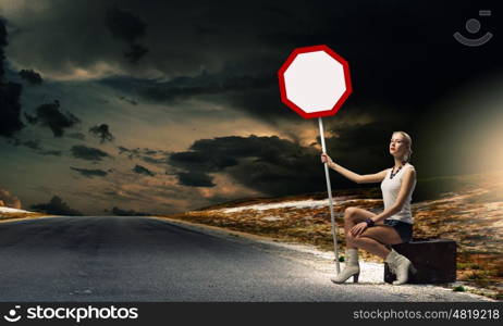 Girl with roadsign. Young woman sitting on suitcase and holding roadsign