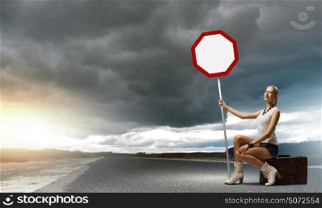 Girl with roadsign. Young woman sitting on suitcase and holding roadsign