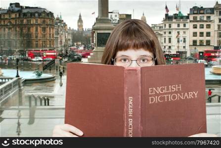 girl with spectacles looks over English Dictionary book and Trafalgar Square in London on background