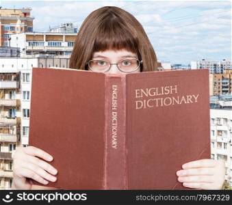 girl with spectacles looks over English Dictionary book with cityscape on background