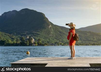 Girl with stalk hat and red dress on taking a selfie by a lake on a dock in front of big mountains. Lifestyle concept