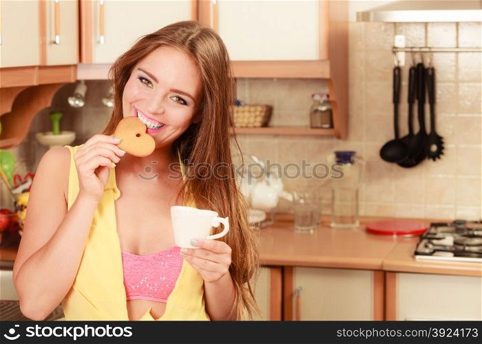 Girl with tea coffee eating gingerbread cookie.. Happy pin up girl with tea or coffee eating heart shape gingerbread cookie. Pretty gorgeous young retro woman with hot beverage relaxing in kitchen.
