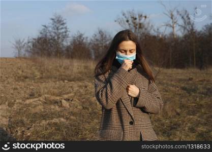 Girl, young European woman in protective sterile medical mask on her face coughs outdoors. Symptoms of virus, Chinese pandemic coronavirus concept., COVID-19, pneumonia, cough. Girl, young European woman in protective sterile medical mask on her face coughs outdoors. Symptoms of virus, Chinese pandemic coronavirus concept., COVID-19, pneumonia, cough.