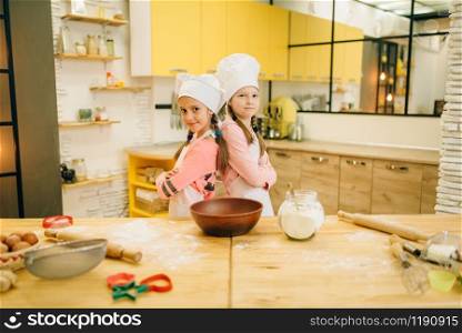 Girls cooks in caps are back to each other on the kitchen. Kids cooking pastry, little chefs holds rolling pin. Girls cooks in caps are back to each other