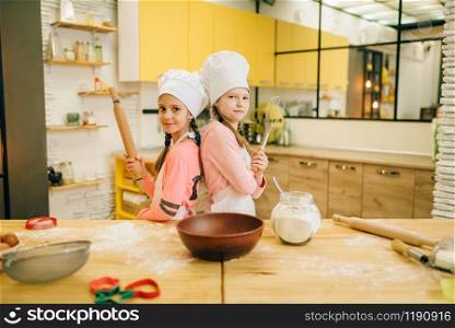 Girls cooks in caps are back to each other on the kitchen. Kids cooking pastry, little chefs holds rolling pin