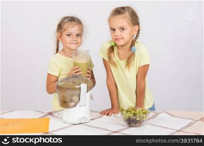 Girls holding a large glass of juice squeezed in a juicer. Two girls at a table squeezed juice from pears and grapes with a juicer