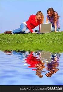 Girls with notebook sitting on grass against sky with reflection on water