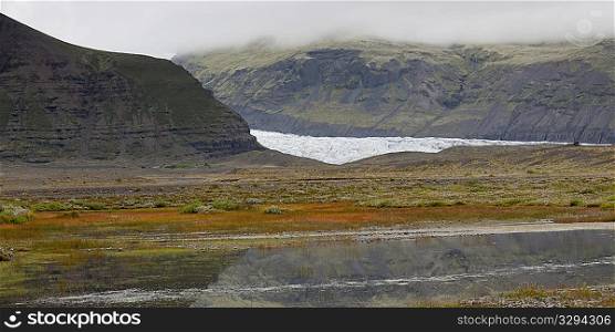 Glacier in misty rugged mountain valley, with water body reflecting the sky