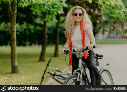 Glad active curly haired female bicyclist covers distance on her own transport, wears sunglasses, casual outfit, poses in park with green trees, expresses good emotions. Riding bicycle concept