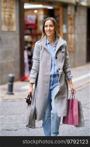 Glad adult woman in stylish outerwear looking away with smile and carrying shopping bags while crossing road in daytime in city. Female shopaholic with paper bags crossing street