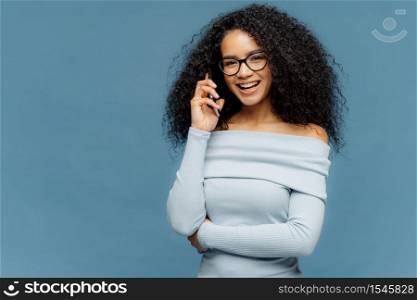 Glad African American female enjoys telephone conversation, keeps one hand over waist, uses modern technologies, wears spectacles and light blue jumper, stands in studio, blank space for your slogan