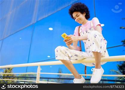 Glad African American female with Afro hairstyle in overall browsing cellphone while sitting on railing near glass building in city. Content black woman texting message on fence