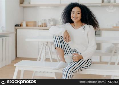 Glad African American woman holds cup of hot beverage, leans at knee, wears white stylish jumper and striped trousers, smiles pleasantly, spends leisure time at home, sits at bench in kitchen