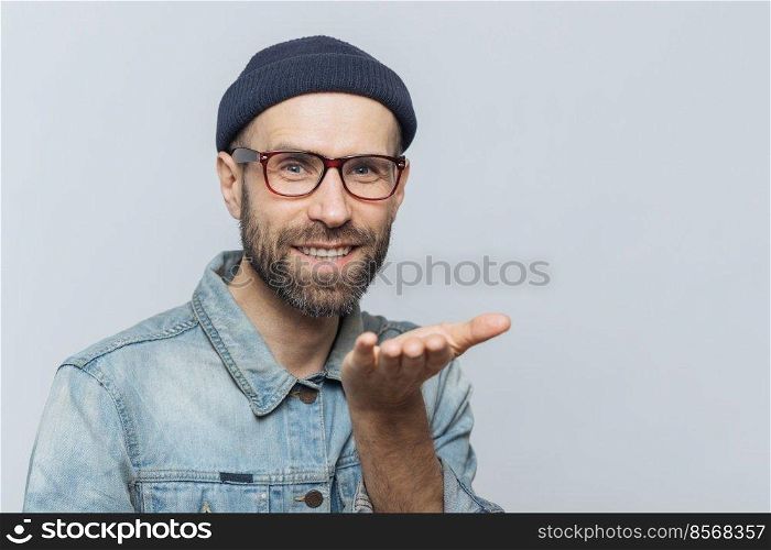 Glad bearded male in trendy eyewear expresses his love, shows air kiss, has cheerful expression, isolated over grey background. Blue eyed middle aged man in stylish clothing, gestures indoor