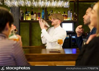 Glad bearded man, in white shirt and cap, smiling and mixing cocktail ingredients in shaker, while standing behind counter near male and female customers in bar. Positive barkeeper preparing cocktail for clients in a pub