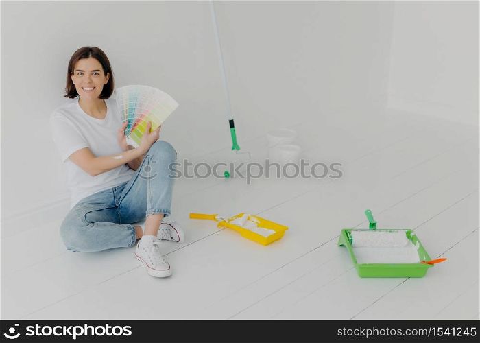 Glad brunette woman demonstrates color samples, dressed in white t shirt and jeans, chooses tone for wall refurbishment, busy with repairing and renovation, looks with smile, thinks about design
