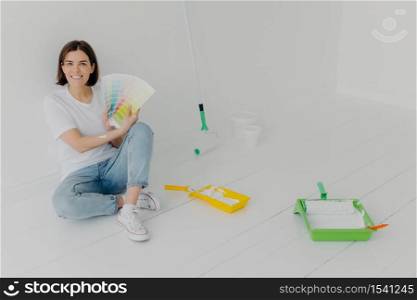 Glad brunette woman demonstrates color samples, dressed in white t shirt and jeans, chooses tone for wall refurbishment, busy with repairing and renovation, looks with smile, thinks about design