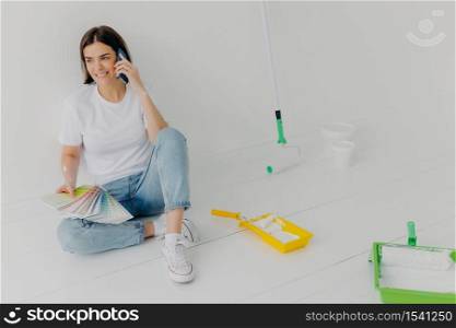 Glad brunette woman in casual clothes, holds color samples, chooses appropriate tone for room, asks for advice in friend, calls via smartphone, poses in new bought apartment, isolated on white wall
