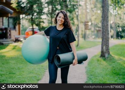 Glad brunette young woman smiles positively holds big fitness ball and karemat, dressed in black t shirt, leggings, going to have gymnastic exercises outdoor, has perfect figure, healthy body