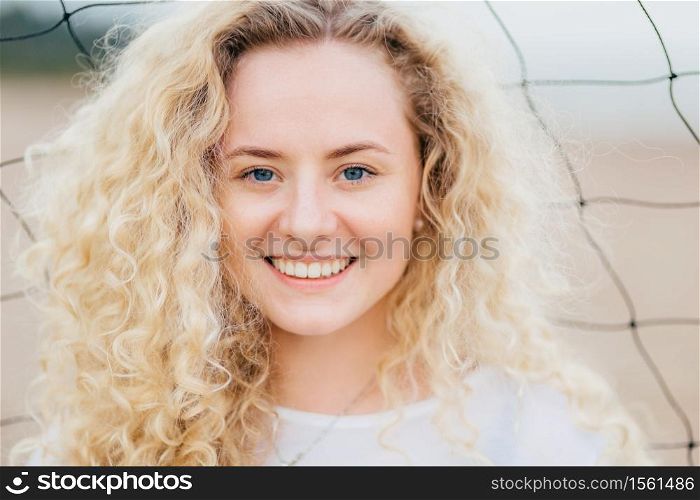 Glad curly Caucasian woman with toothy smile, has curly bushy light hair, blue eyes, clean skin, stands outdoor, has appealing appearance. Close up portrait of good looking female rests at beach
