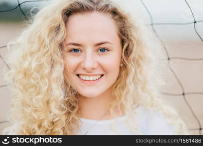 Glad curly Caucasian woman with toothy smile, has curly bushy light hair, blue eyes, clean skin, stands outdoor, has appealing appearance. Close up portrait of good looking female rests at beach