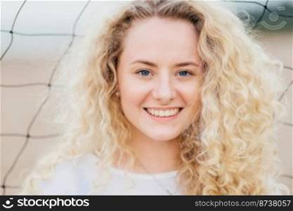 Glad curly Caucasian woman with toothy smile, has curly bushy light hair, blue eyes, clean skin, stands outdoor, has appealing appearance. Close up portrait of good looking female rests at beach