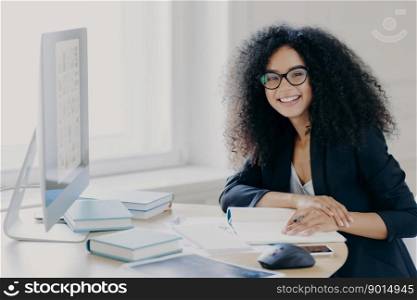 Glad curly office worker watches webinar on business topic, writes down notes with pen, smiles positively, wears spectacles and black clothes prepares report wears transparent glasses. Coworking space