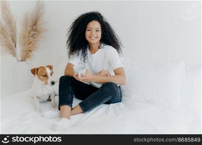 Glad dark skinned Afro American female feels relaxed, poses in bedroom on comfortable bed with pedigree dog, drinks hot beverage, has morning coffee, smiles happily, enjoys spare time at home.