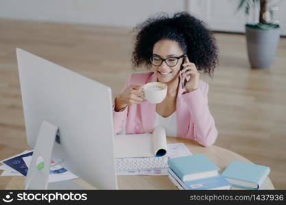 Glad dark skinned business lady looks happily at computer, drinks fresh hot beverage, holds modern mobile phone, dressed elegantly, poses against office interior, sits at desk with textbook, notepad