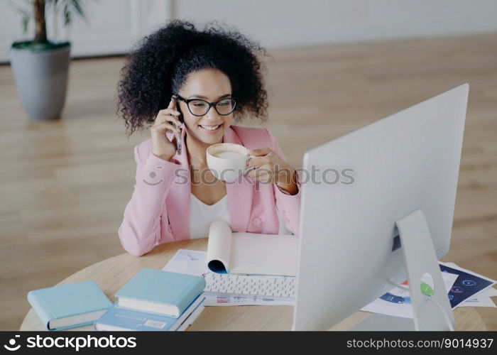 Glad dark skinned business lady looks happily at computer, drinks fresh hot beverage, holds modern mobile phone, dressed elegantly, poses against office interior, sits at desk with textbook, notepad