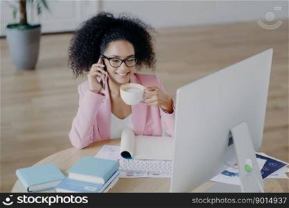 Glad dark skinned business lady looks happily at computer, drinks fresh hot beverage, holds modern mobile phone, dressed elegantly, poses against office interior, sits at desk with textbook, notepad
