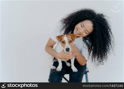 Glad dark skinned girl plays with jack russell terrier dog, have fun together, poses against white background, dressed casually. Happy Afro girl poses with pet indoor. People, animals, fun concept