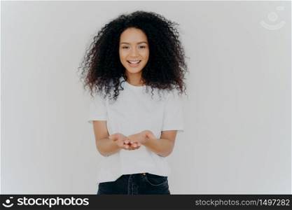 Glad dark skinned woman has cupped hands, asks to give something, smiles plesantly, has bushy frizzy hair, wears t shirt and jeans, isolated over white background. People and charity concept
