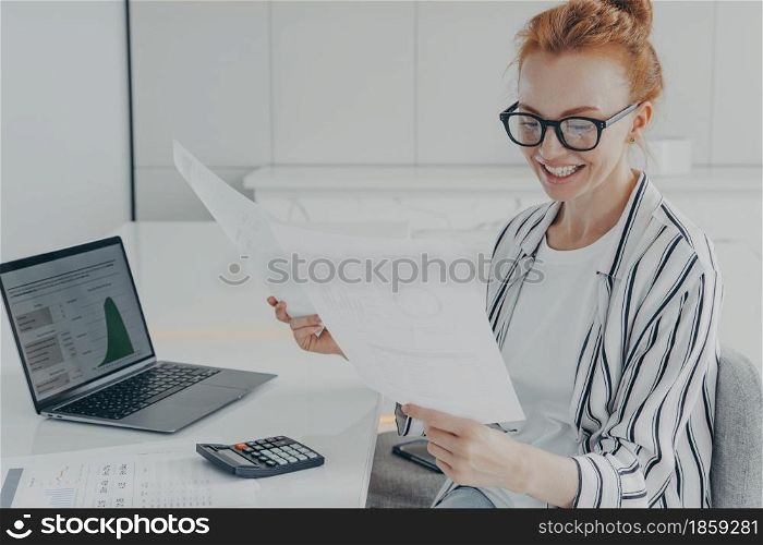 Glad European female financier studies financial graphs diagrams counts expenses holds paper documents sits at desktop with laptopn computer and calculator wors at home office manages budget. Glad European female financier studies financial graphs diagrams counts expenses holds papers