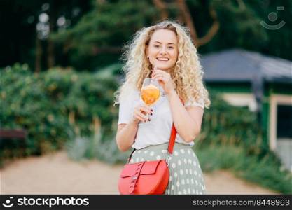 Glad European female with curly blonde hair, wears casual clothes, holds red stylish bag, drinks fresh summer cocktail, walks across park, looks joyfully into distance stands outdoor. Leisure concept