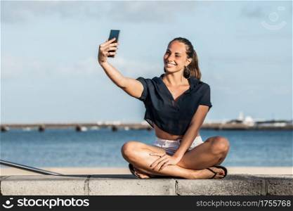 Glad female sitting on stone fence on seashore and taking self shot on smartphone while enjoying weekend on Lanzarote in summer. Smiling woman taking selfie on embankment