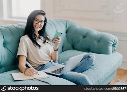 Glad female student works on course paper, surrounded with modern gadget, makes notes in notepad, feels relaxed at comfortable sofa, works online, wears casual clothes, watches video online.