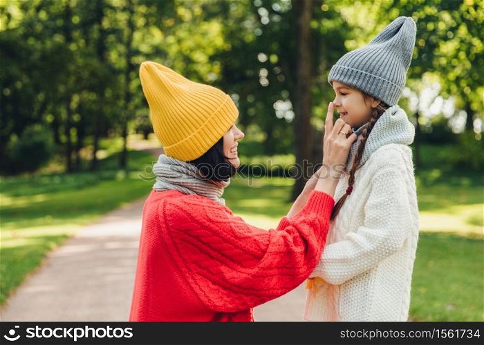 Glad female wears trendy yellow hat and red sweater with scarf touches nose of her small daughter, have walk together in park, play and communicate. People, relationship and togetherness concept